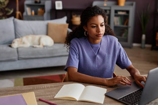 A woman uses a laptop as a dog sits on a couch behind her