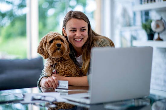 A person holds a dog and credit card while sitting in front of a laptop
