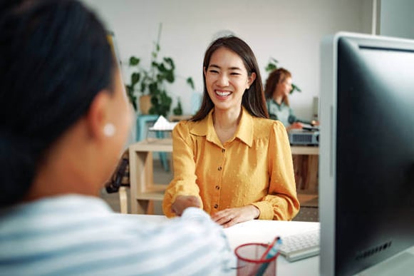 A woman sits at a desk shaking another woman's hand