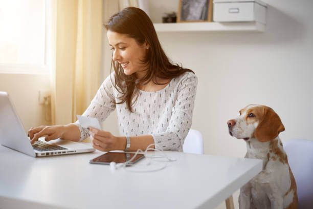 A woman holds a credit card while using a laptop as a dog sits next to her